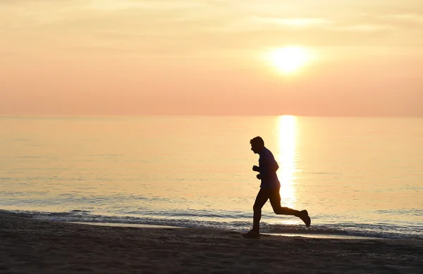 Silhueta jovem homem do esporte correndo ao ar livre na praia ao pôr do sol com céu laranja — Fotografia de Stock