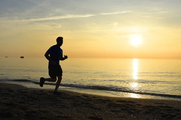 Siluet pemuda olahraga berjalan di luar rumah di pantai saat matahari terbenam dengan langit oranye — Stok Foto