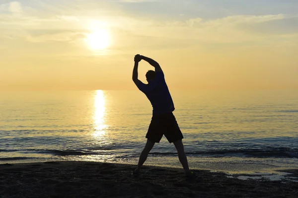 Silhouette del giovane sportivo allungamento gamba dopo l'allenamento all'aperto sulla spiaggia al tramonto — Foto Stock