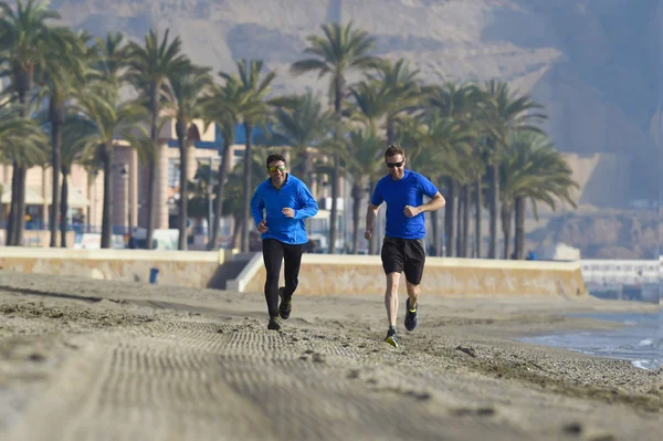 Dos hombres amigos corriendo juntos en playa arena con palmeras fondo en sesión de entrenamiento de la mañana trotando entrenamiento uno en manga larga y pantalones el otro chico en pantalones cortos — Foto de Stock