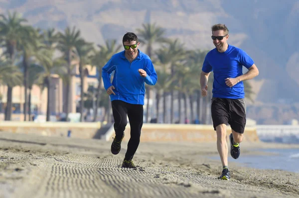 Dos hombres amigos corriendo juntos en playa arena con palmeras fondo en sesión de entrenamiento de la mañana trotando entrenamiento uno en manga larga y pantalones el otro chico en pantalones cortos — Foto de Stock