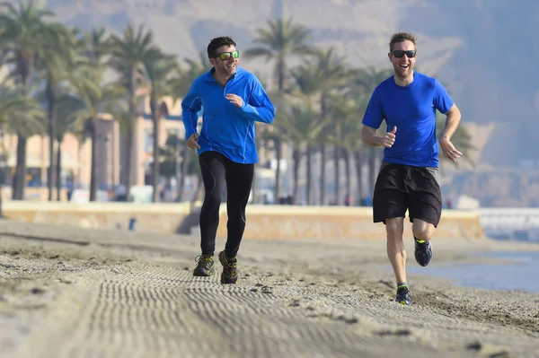 Dos hombres amigos corriendo juntos en playa arena con palmeras fondo en sesión de entrenamiento de la mañana trotando entrenamiento uno en manga larga y pantalones el otro chico en pantalones cortos — Foto de Stock