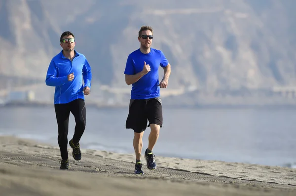 two men friends running together on beach sand with beautiful coast mountain background in morning training session jogging workout one in long sleeve and pants the other guy in shorts