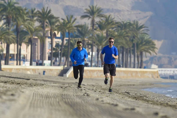 Dos hombres amigos corriendo juntos en playa arena con palmeras fondo en sesión de entrenamiento de la mañana trotando entrenamiento uno en manga larga y pantalones el otro chico en pantalones cortos — Foto de Stock