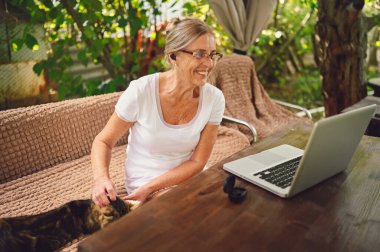 Teknoloji, yaşlılar konsepti. Ev kedisi olan yaşlı mutlu bir kadın bahçede dizüstü bilgisayarla çalışan kablosuz kulaklık kullanıyor. Uzak çalışma, uzak eğitim. 