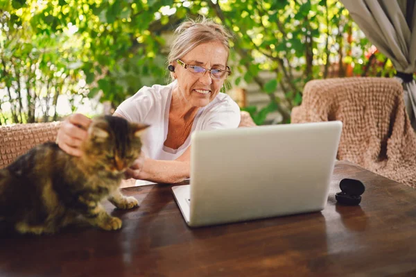 Technology, old age people concept - elderly happy senior woman with home cat use wireless headphones working online with laptop computer outdoor in the garden. Remote work, distance education.