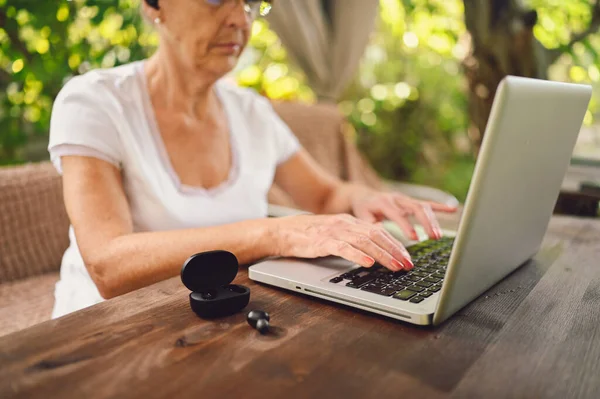 Technology, old age people concept - elderly senior old woman hands working online with laptop computer outdoor in the garden. Wireless headphones in the foreground. Remote work, distance education.