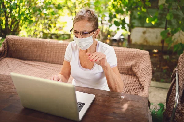 Technology, old age people concept - elderly senior woman in protective face mask use wireless headphones working online with laptop computer outdoor in the garden. Remote work, distance education.