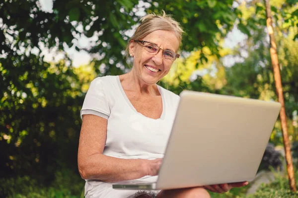 Technology, old age people concept - elderly happy senior old woman working online with laptop computer outdoor in the garden. Remote work, distance education.