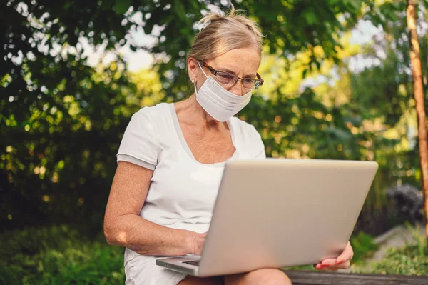 Technology, old age people concept - elderly senior old woman in face protective mask working online with laptop computer outdoor in the garden. Remote work, distance education.