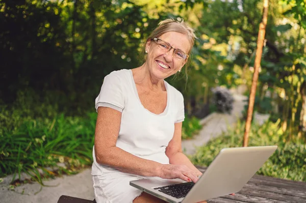 Technology, old age people concept - elderly happy senior old woman working online with laptop computer outdoor in the garden. Remote work, distance education.