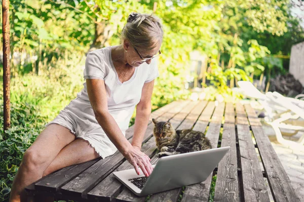 Wanita tua yang bahagia dengan kucing rumahan yang bekerja secara online dengan komputer laptop di luar — Stok Foto