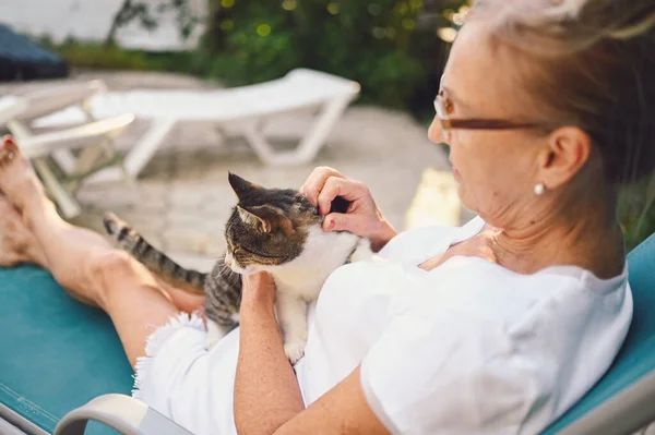 Happy smiling senior elderly woman in glasses relaxing in summer garden outdoors hugging domestic tabby cat. Retired old people and animals pets concept