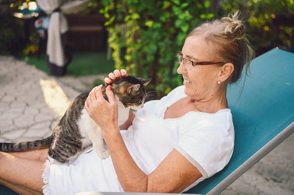 Happy smiling senior elderly woman in glasses relaxing in summer garden outdoors hugging domestic tabby cat. Retired old people and animals pets concept