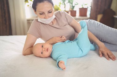 Koruyucu yüz maskesi takmış genç bir anne, mavi tulumlu şirin bir bebek, onu yatakta kucaklıyor. Sağlıklı çocuk, hastane kavramı ve mutlu annelik. Çocuk odası..