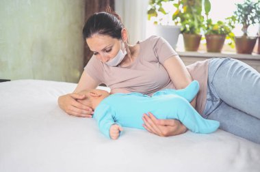 Koruyucu yüz maskesi takmış genç bir anne mavi tulumlu şirin bir bebekle onu kucaklıyor ve anne sütüyle besliyor. Sağlıklı çocuk, hastane kavramı ve mutlu annelik. Çocuk odası..