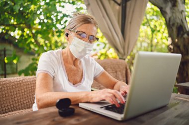 Teknoloji, yaşlılar konsepti - koruyucu yüz maskeli yaşlı kadın bahçede dizüstü bilgisayarla çalışan kablosuz kulaklık kullanıyor. Uzak çalışma, uzak eğitim. 