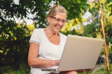 Teknoloji, yaşlılar konsepti. Yaşlı mutlu yaşlı kadın bahçede dizüstü bilgisayarla çalışıyor. Uzak çalışma, uzak eğitim. 