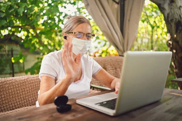 Technology, old age people concept - elderly senior woman in protective face mask use wireless headphones working online with laptop computer outdoor in the garden. Remote work, distance education.