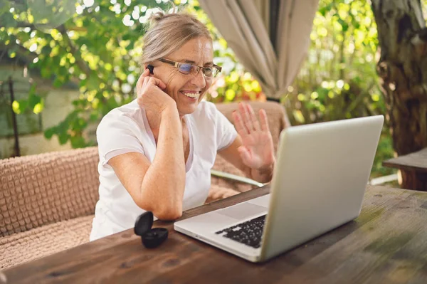 Technology, old age people concept - elderly happy senior woman using wireless headphones working online with laptop computer outdoor in the garden. Remote work, distance education.