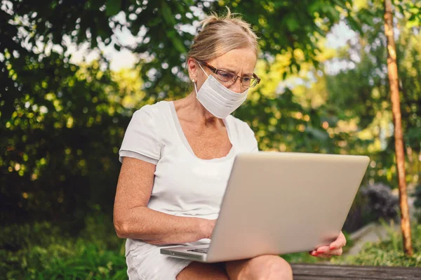 Technology, old age people concept - elderly senior old woman in face protective mask working online with laptop computer outdoor in the garden. Remote work, distance education.