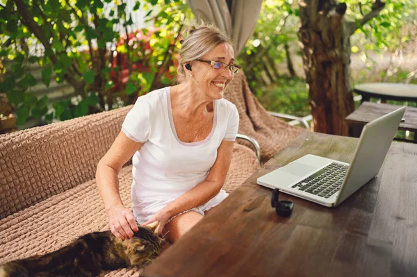 Technology, old age people concept - elderly happy senior woman with home cat use wireless headphones working online with laptop computer outdoor in the garden. Remote work, distance education.