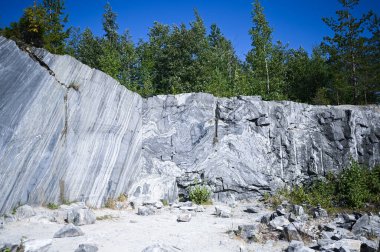 Kaya, ağaçlar ve mavi gökyüzü ile doğal ortamda beyaz ve gri mermerden oluşan ham bir yüzeyi kesti. Mavi mermer kariyer dokusu. Arkaplan taşı, binalar ve iç mekanlar için endüstriyel malzeme.
