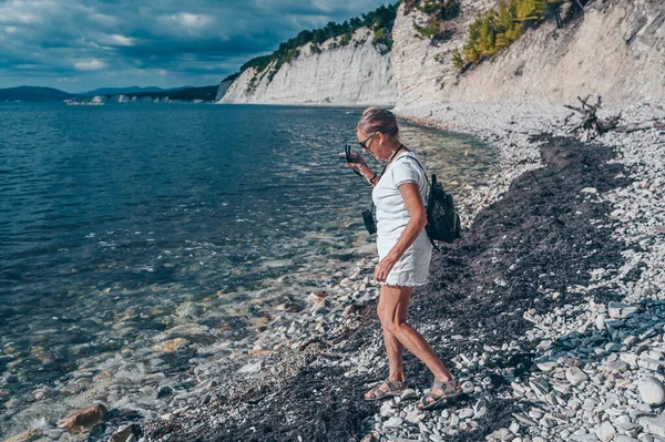 นักท่องเที่ยวหญิงผู้สูงอายุเดินทางกระเป๋าเป้สะพายหลังผู้สูงอายุ ถ่ายรูปชายฝั่งทะเล หิน ท้องฟ้าสีฟ้า — ภาพถ่ายสต็อก