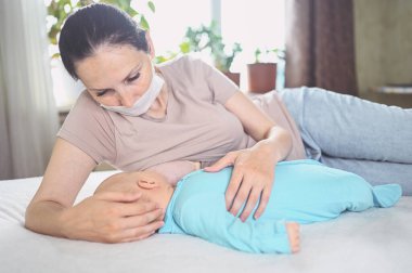 Koruyucu yüz maskesi takmış genç bir anne mavi tulumlu şirin bir bebekle onu kucaklıyor ve anne sütüyle besliyor. Sağlıklı çocuk, hastane kavramı ve mutlu annelik. Çocuk odası..
