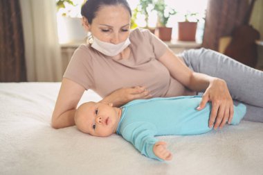 Koruyucu yüz maskesi takmış genç bir anne, mavi tulumlu şirin bir bebek, onu yatakta kucaklıyor. Sağlıklı çocuk, hastane kavramı ve mutlu annelik. Çocuk odası..