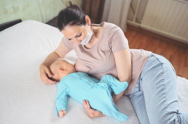 Koruyucu yüz maskesi takmış genç bir anne mavi tulumlu şirin bir bebekle onu kucaklıyor ve anne sütüyle besliyor. Sağlıklı çocuk, hastane kavramı ve mutlu annelik. Çocuk odası..