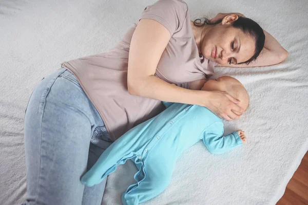 stock image Young mother lies with a newborn cute infant baby in blue jumpsuit on the bed, hugging and breastfeeds him with breast milk. Healthy child, concept of hospital and happy motherhood. Nursery.