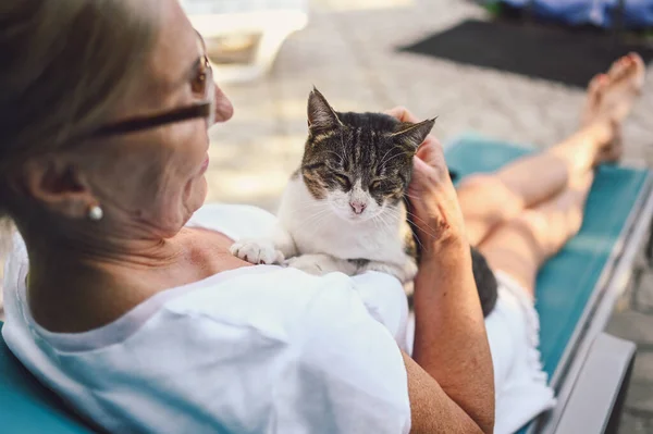 Happy smiling senior elderly woman in glasses relaxing in summer garden outdoors hugging domestic tabby cat. Retired old people and animals pets concept