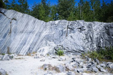 Kaya, ağaçlar ve mavi gökyüzü ile doğal ortamda beyaz ve gri mermerden oluşan ham bir yüzeyi kesti. Mavi mermer kariyer dokusu. Arkaplan taşı, binalar ve iç mekanlar için endüstriyel malzeme.