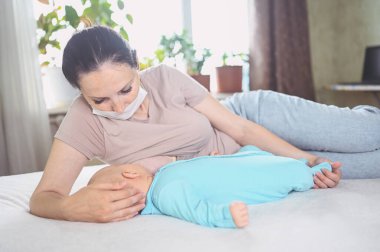 Koruyucu yüz maskesi takmış genç bir anne mavi tulumlu şirin bir bebekle onu kucaklıyor ve anne sütüyle besliyor. Sağlıklı çocuk, hastane kavramı ve mutlu annelik. Çocuk odası..