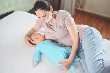 Koruyucu yüz maskesi takmış genç bir anne mavi tulumlu şirin bir bebekle onu kucaklıyor ve anne sütüyle besliyor. Sağlıklı çocuk, hastane kavramı ve mutlu annelik. Çocuk odası..