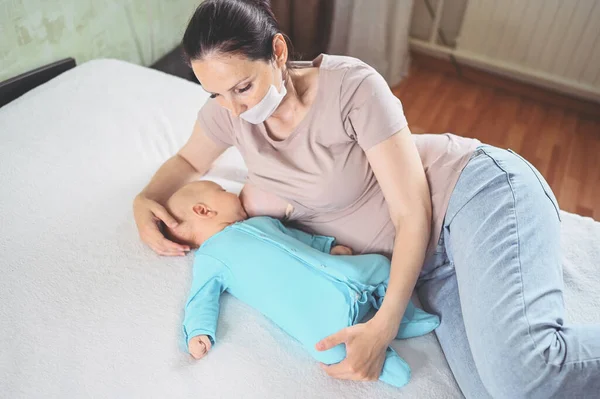 Koruyucu yüz maskesi takmış genç bir anne mavi tulumlu şirin bir bebekle onu kucaklıyor ve anne sütüyle besliyor. Sağlıklı çocuk, hastane kavramı ve mutlu annelik. Çocuk odası..