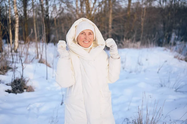 Happy elderly senior mature woman in white warm outwear playing with snow in sunny winter outdoors. Retired healthy people holiday vacation winter activities, active lifestyle concept