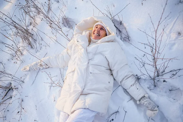 Happy elderly senior mature woman in white warm outwear playing with snow in sunny winter outdoors. Retired healthy people holiday vacation winter activities, active lifestyle concept