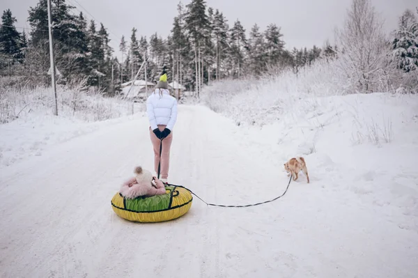 外を歩くピンク色の暖かい外で幸せな母親と小さなかわいい女の子雪の白い寒い冬の森の中で赤い柴犬の犬と楽しい乗り物インフレータブル雪チューブを屋外で持っています 家族スポーツ休暇活動 — ストック写真