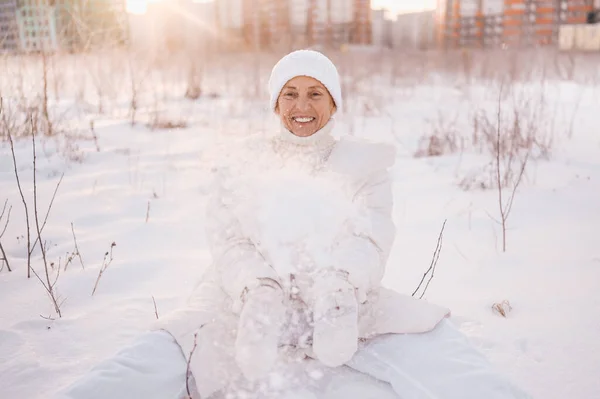 Happy elderly senior mature woman in white warm outwear playing with snow in sunny winter outdoors. Retired healthy people holiday vacation winter activities, active lifestyle concept