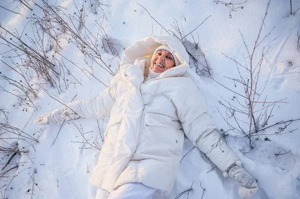 Happy elderly senior mature woman in white warm outwear playing with snow in sunny winter outdoors. Retired healthy people holiday vacation winter activities, active lifestyle concept