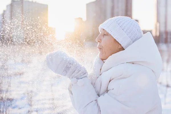 Happy elderly senior mature woman in white warm outwear playing with snow in sunny winter outdoors. Retired healthy people holiday vacation winter activities, active lifestyle concept