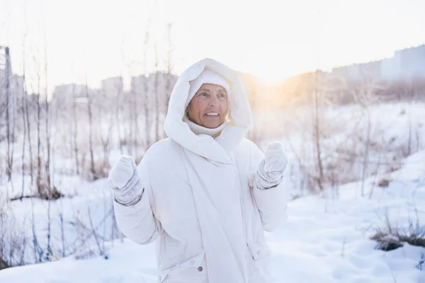 Happy elderly senior mature woman in white warm outwear playing with snow in sunny winter outdoors. Retired healthy people holiday vacation winter activities, active lifestyle concept