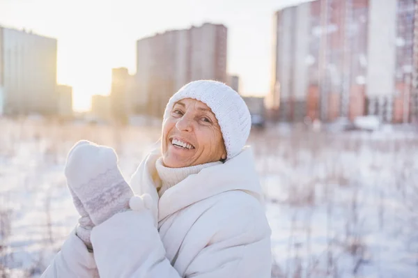Happy elderly senior mature woman in white warm outwear playing with snow in sunny winter outdoors. Retired healthy people holiday vacation winter activities, active lifestyle concept