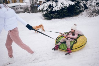 Mutlu anne ve küçük sevimli kız pembe, sıcak, yürüyüş kıyafetleri içinde, kar yağışı gibi beyaz kış ormanlarında kırmızı shiba inu köpeğiyle şişirilebilir kar tüpü sürerken eğleniyorlar. Aile spor tatili aktiviteleri