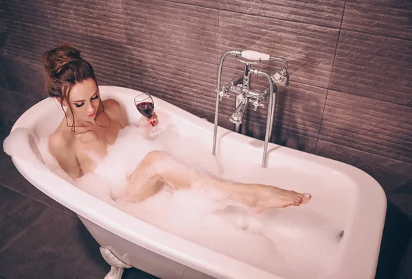 漂亮的年轻女子化妆和发型与一杯红酒放松沐浴在一个充满泡沫的复古浴缸 温泉美容院的概念 身体和皮肤护理 — 图库照片