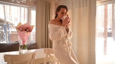 İnce, güzel bir kadın, uzun saçlı, düşük beyaz bornozlu, güneşli bir sabah geçiriyor, İskandinav tarzı bir kır evinin oturma odasında çay ya da kahve içiyor. İnsanlar, güzellik, vücut bakım spa konsepti