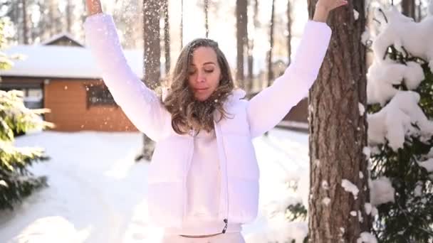 雪に覆われた緑のトウヒの木の背景に田舎の木造住宅の裏庭に白い暖かい外耳道のジャケットで美しい若い女性の雪の中で冬の日当たりの良い冷たい肖像画 — ストック動画
