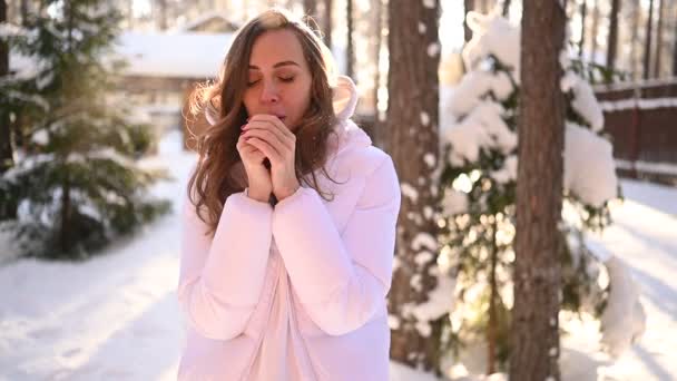 雪に覆われた緑のトウヒの木の背景に田舎の木造住宅の裏庭に白い暖かい外耳道のジャケットで美しい若い女性の雪の中で冬の日当たりの良い冷たい肖像画 — ストック動画